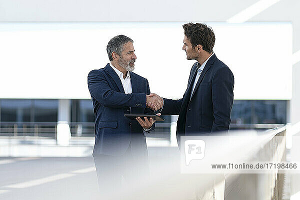 Mature entrepreneur holding digital tablet while doing handshake with colleague standing at office building terrace