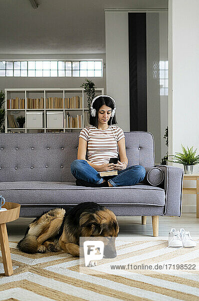 Woman listening to music sitting on sofa while dog sleeping on carpet in living room
