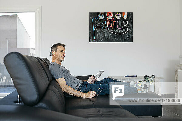 Mature man with digital tablet looking away while sitting on sofa at home