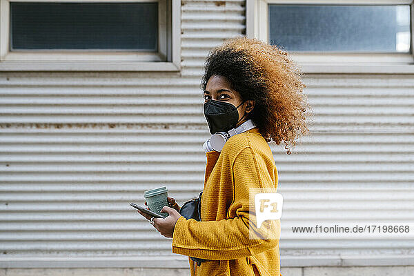 Junge Frau mit Kaffeetasse und Handy in der Hand  die während einer Pandemie an der Wand steht