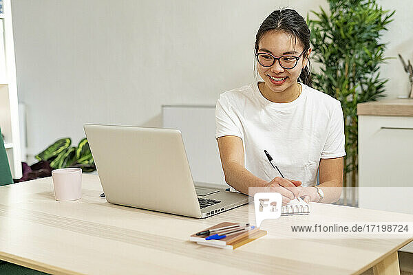 Smiling woman writing in notepad while sitting with laptop at home