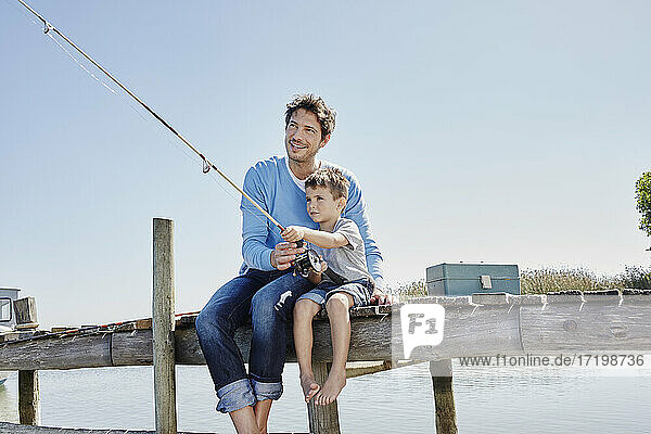 Lächelnder Mann  der einem Jungen beim Angeln hilft  während er auf dem Pier sitzt