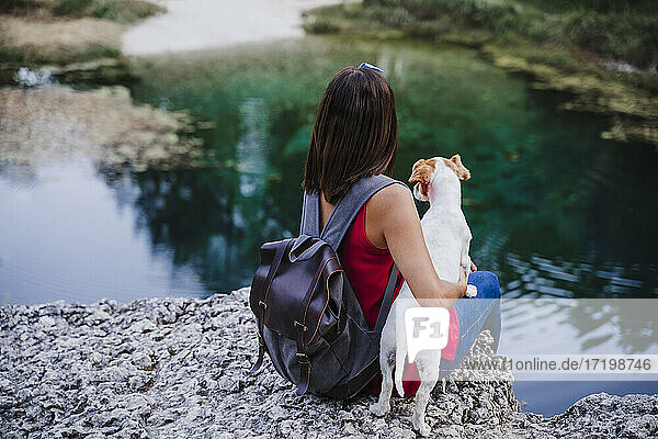 Woman with backpack embracing dog while sitting on rock at lake