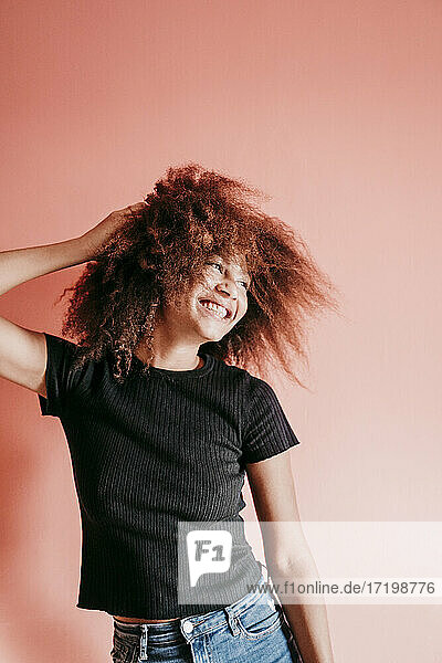 Fröhliche Afro-Frau mit Hand im Haar  die vor einem pfirsichfarbenen Hintergrund wegschaut
