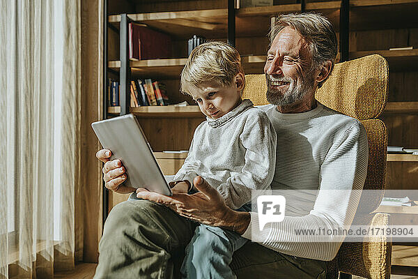 Junge  der auf dem Schoß seines lächelnden Vaters im Wohnzimmer sitzt und e-learning betreibt