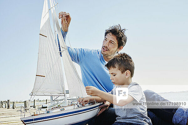 Vater und Sohn bereiten gemeinsam ein Spielzeug-Segelboot vor  während sie am Pier sitzen