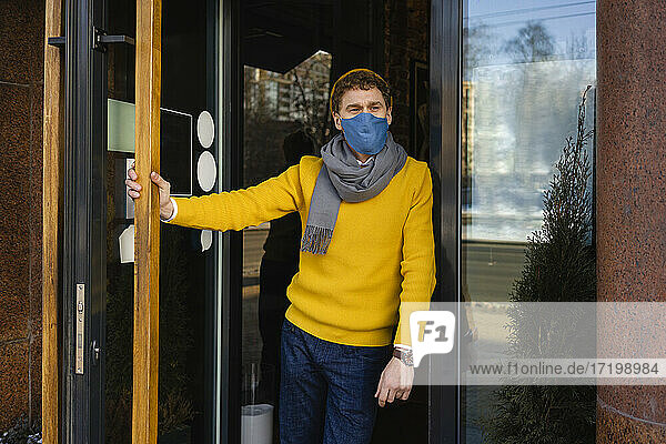Male owner wearing protective face mask leaning at cafe entrance during COVID-19
