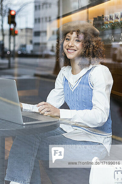 Teenager-Mädchen mit Baskenmütze sitzt lächelnd am Laptop in einem Cafe