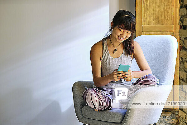 Smiling woman using mobile phone while sitting on arm chair at home
