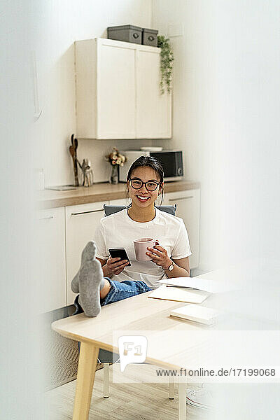Frau mit Mobiltelefon und Kaffeetasse lächelnd beim Entspannen am Tisch zu Hause