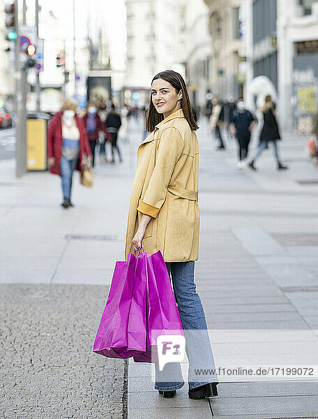Lächelnde junge Frau mit magentafarbenen Einkaufstaschen auf dem Fußweg in der Stadt