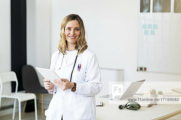 Mittlere erwachsene Ärztin mit digitalem Tablet  die sich im Stehen auf den Schreibtisch stützt