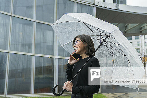 Fröhliche junge Frau  die einen Regenschirm hält  während sie bei Regen an einem Glasgebäude telefoniert