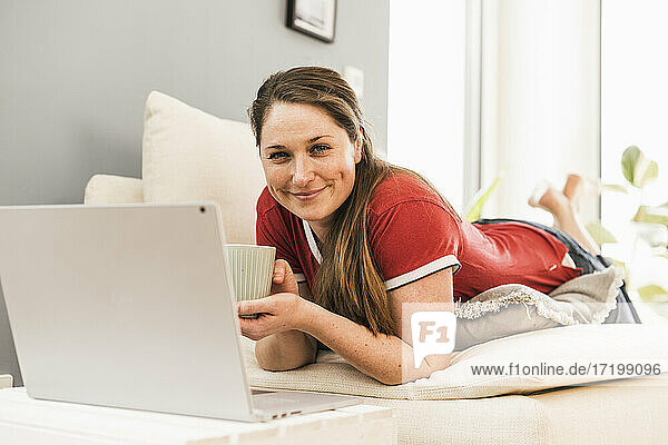 Lächelnde Frau mit Kaffeetasse auf dem Sofa zu Hause liegend