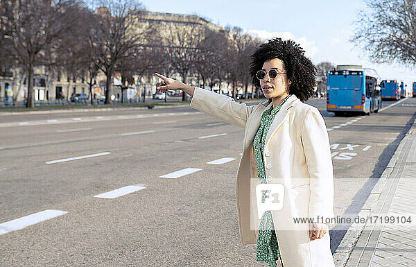 Junge Frau mit Sonnenbrille  die auf einem Gehweg in der Stadt steht und eine Mitfahrgelegenheit sucht