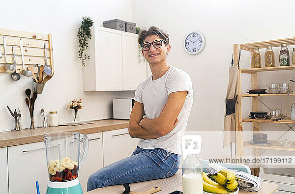 Lächelnder junger Mann sitzt mit verschränkten Armen auf einem Tisch in der Küche