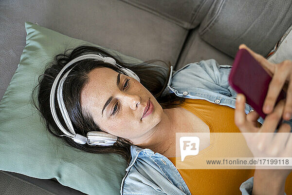 Junge Frau mit Kopfhörern  die ein Mobiltelefon benutzt  während sie zu Hause auf dem Sofa liegt