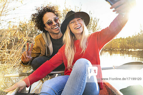 Lächelnde Frau  die ein Selfie mit einem Mann durch ein Mobiltelefon macht  während sie in einem Kanu auf einem Fluss sitzt