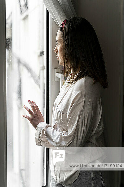 Nachdenkliche junge Frau schaut durch das Fenster  während sie zu Hause steht