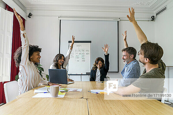 Cheerful male and female entrepreneurs with arms raised in board room at office