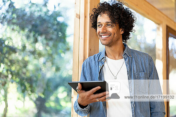 Lächelnder junger Mann  der ein digitales Tablet hält  während er am Fenster im Vorgarten steht