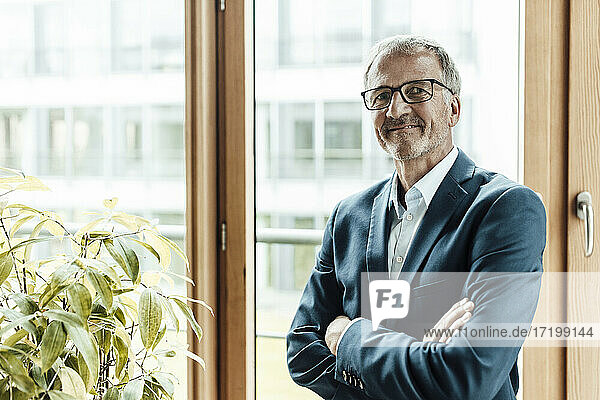 Smiling businessman with arms crossed standing at glass window in office