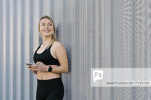 Lächelnde Sportlerin in Sportkleidung  die wegschaut und ihr Handy an die Wand hält