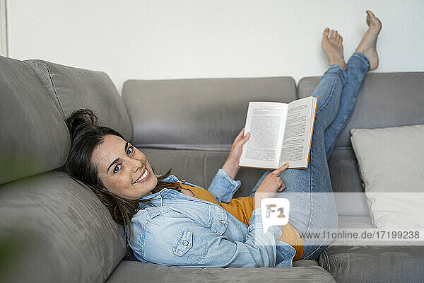 Lächelnde Frau mit Buch auf Sofa liegend