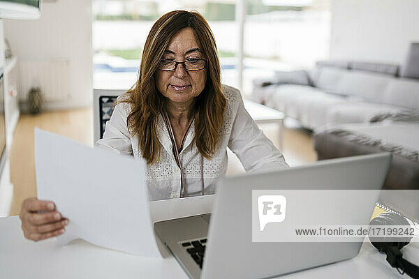 Unternehmerin hält Papier in der Hand  während sie zu Hause einen Laptop benutzt
