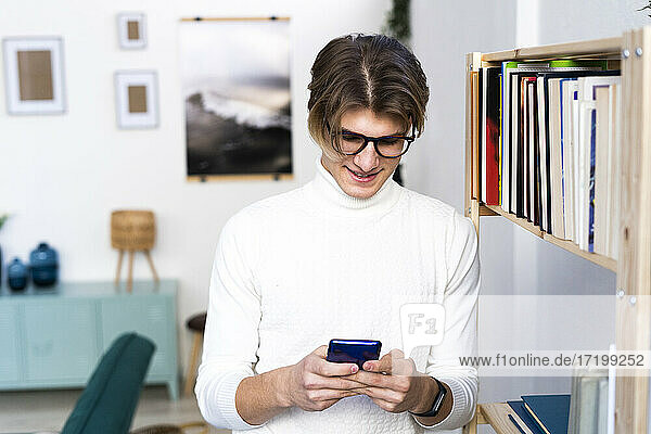 Junger Mann schreibt SMS über sein Smartphone  während er im Wohnzimmer neben einem Bücherregal steht