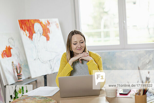 Lächelnde Frau mit Laptop auf dem Tisch  während sie im Heimstudio sitzt