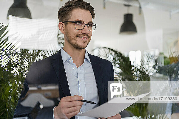 Lächelnder männlicher Unternehmer mit Dokument durch Glas im Büro gesehen