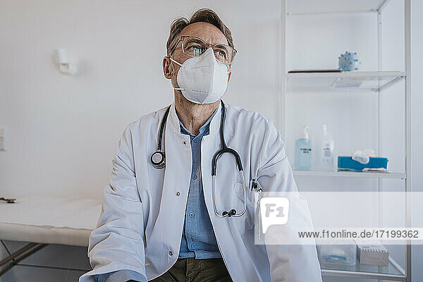 Nachdenklicher Arzt mit Gesichtsschutzmaske  der wegschaut  während er in der Klinik sitzt