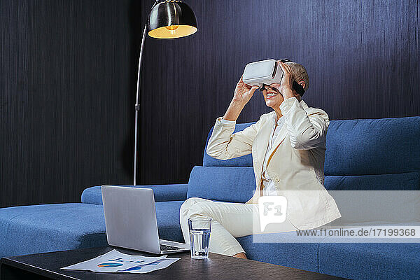 Lächelnde Geschäftsfrau  die ein Virtual-Reality-Headset benutzt  während sie mit einem Laptop auf dem Sofa sitzt