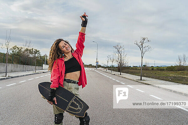 Lächelnde Frau mit Skateboard gestikuliert Horn Zeichen  während auf der Straße gegen den Himmel stehen