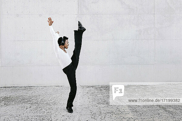 Flexible male dancer stretching leg while practicing urban dance against white wall