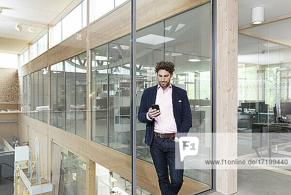 Businessman using smart phone while leaning on glass wall at factory
