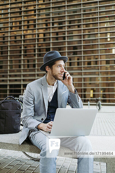 Male entrepreneur wearing hat sitting with laptop on bench while talking on smart phone