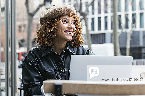 Lächelndes Teenager-Mädchen mit Baskenmütze sitzt mit Laptop in einem Straßencafé