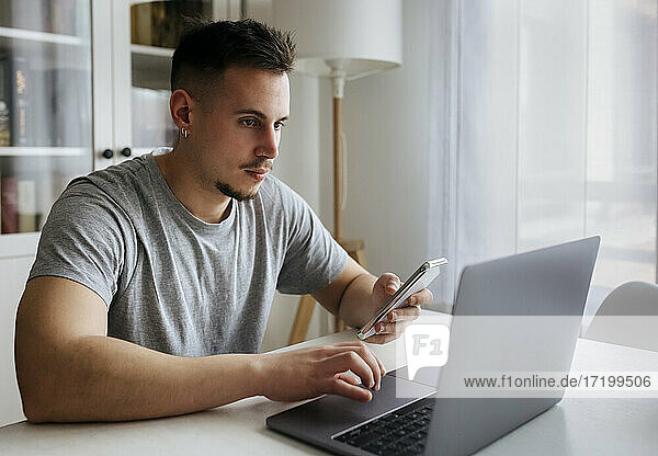 Handsome male entrepreneur with smart phone using laptop on table at home office