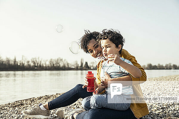 Boy playing with bubble wand while sitting on mother's lap during sunny day