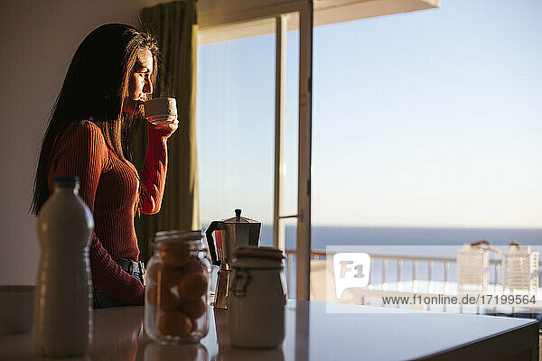 Nachdenkliche Frau  die einen Kaffee trinkt und durch ein Fenster in ihrem Haus schaut