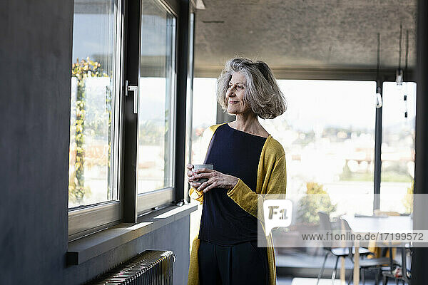 Smiling woman with coffee cup looking through window while standing at home