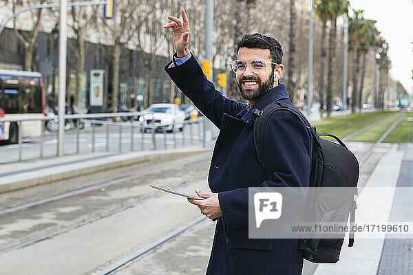 Smiling young businessman with digital tablet waving while standing at station