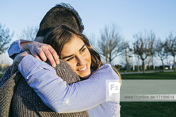 Freundin lächelt  während sie ihren Freund im Park bei Sonnenuntergang umarmt