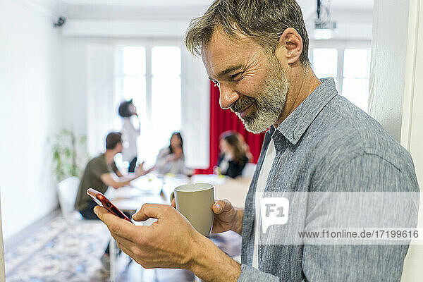 Lächelnder männlicher Unternehmer  der während einer Kaffeepause im Büro ein Smartphone benutzt