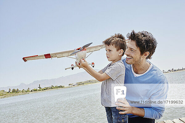 Junge hält Flugzeugspielzeug und steht neben seinem Vater an einem sonnigen Tag