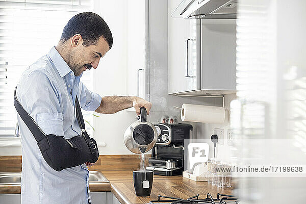 Verletzter Mann mit Armschlinge gießt Tee in Tasse durch Wasserkocher in Küche zu Hause