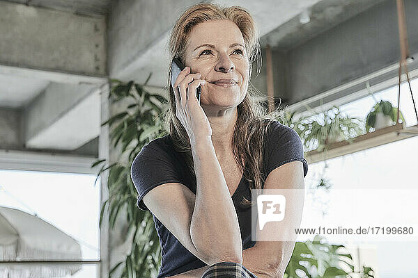 Lächelnde Frau  die wegschaut  während sie in einer Dachgeschosswohnung zu Hause mit dem Handy telefoniert