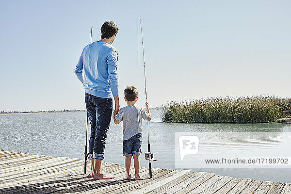 Father and son holding hands while standing with fishing rods on jetty